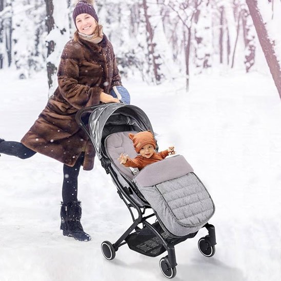 Maman souriante promenant son bébé dans la neige, installé confortablement dans une chancelière grise d'hiver.