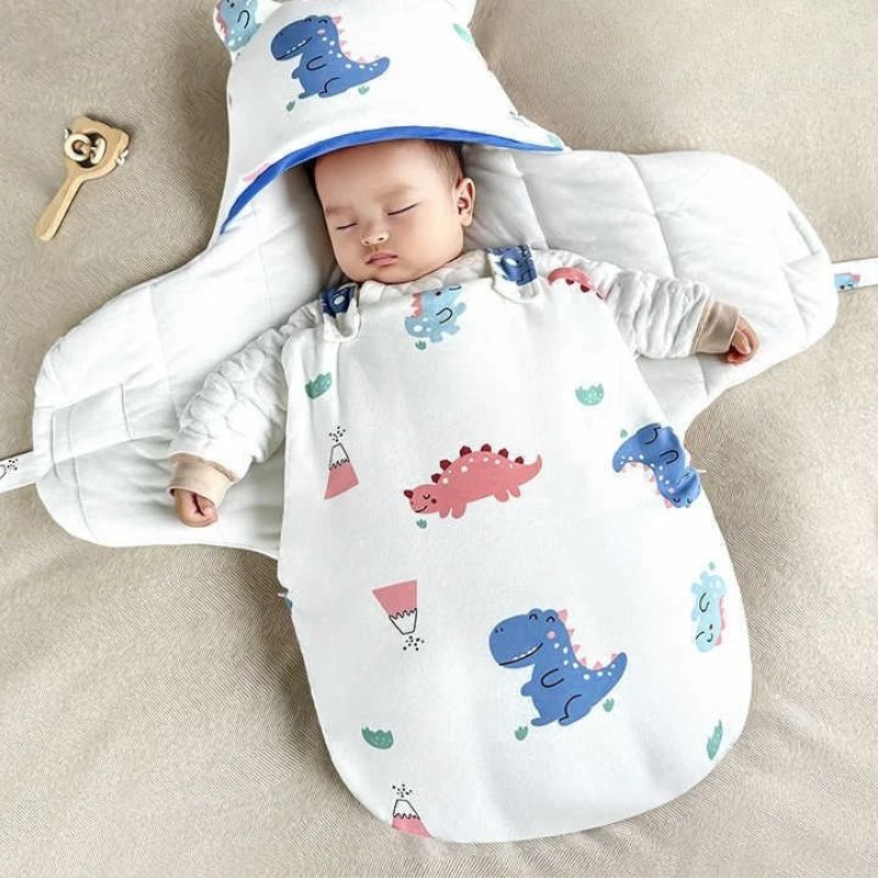 Bébé dormant dans un nid d'ange ouvert à motif dinosaures bleus et rose, sur un lit doux beige.