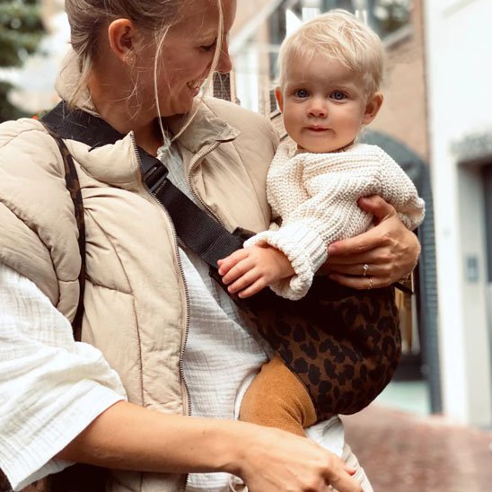 Maman souriante portant son bébé avec une écharpe bébé motif léopard pendant une promenade automnale.
