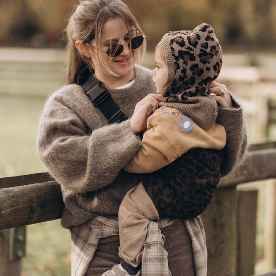 Maman souriante portant son bébé avec une écharpe de portage bébé motif léopard, promenade en automne.