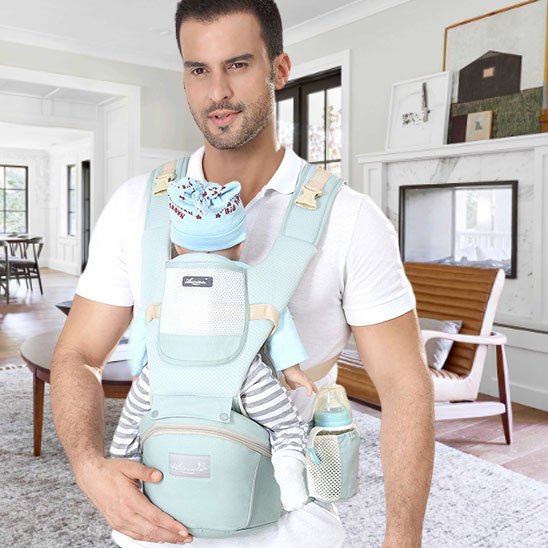 Papa portant son bébé dans un porte-bébé ergonomique bleu clair, appui ventral pratique et sécurisé à la maison.